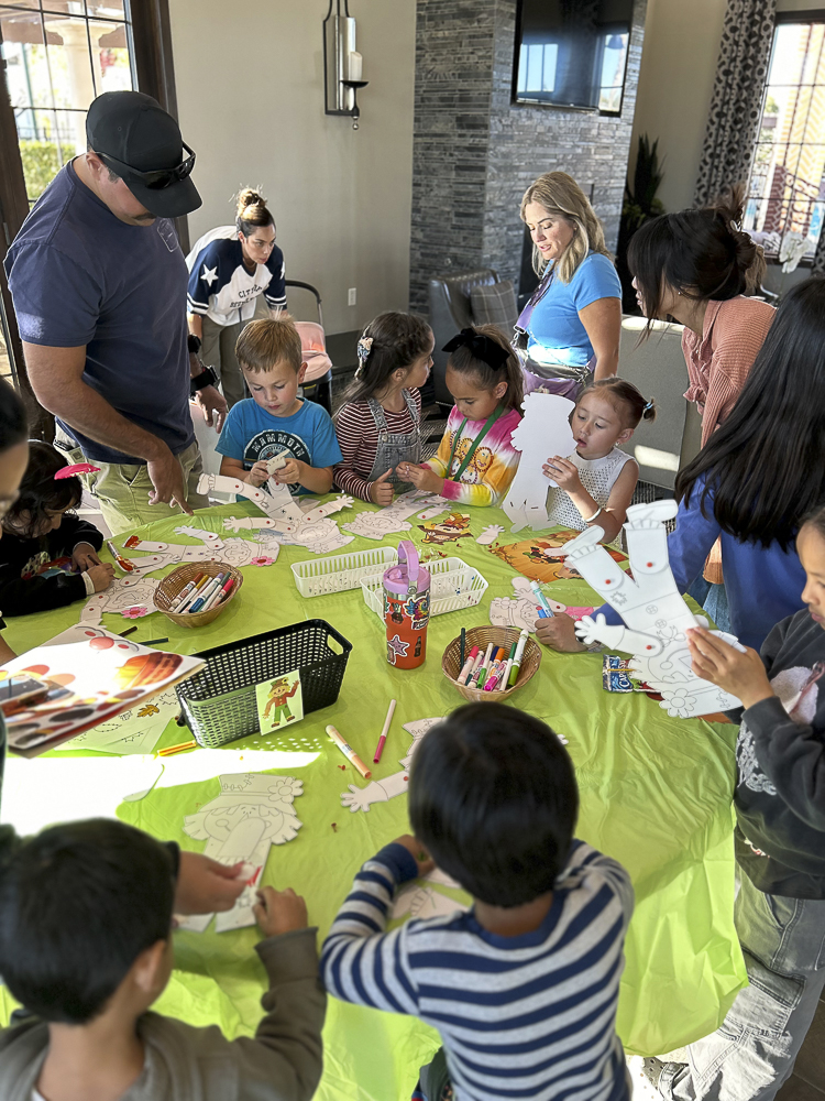 Children and adults engage in crafts at a table covered with a bright green cloth. The kids are coloring and cutting paper shapes, surrounded by markers and supplies, while adults assist and observe the creative activities.