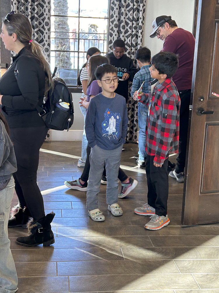 A group of children and adults gather indoors. Two boys in the foreground are engaged in conversation, one wearing glasses and a space-themed shirt, the other in a red plaid shirt. The room has wooden flooring and patterned curtains.