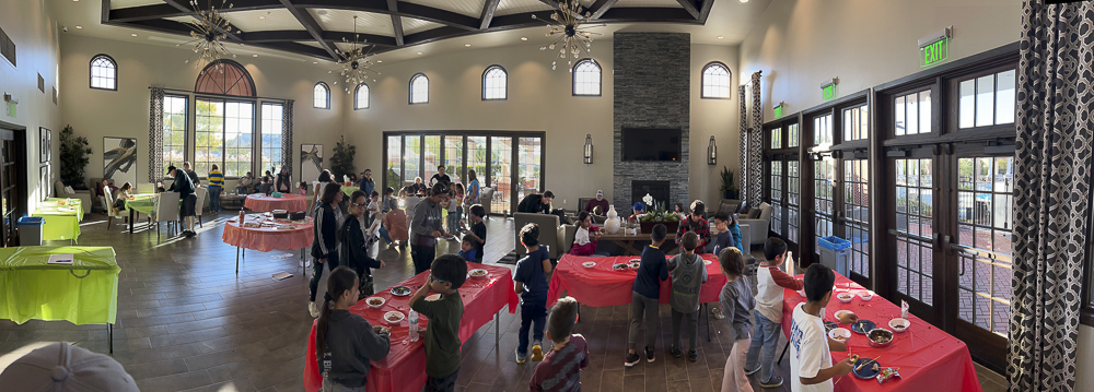 A lively indoor gathering with people of various ages working on crafts at tables covered with red and pink tablecloths. The room is spacious with large windows, high ceilings, and a fireplace. The atmosphere appears social and relaxed.