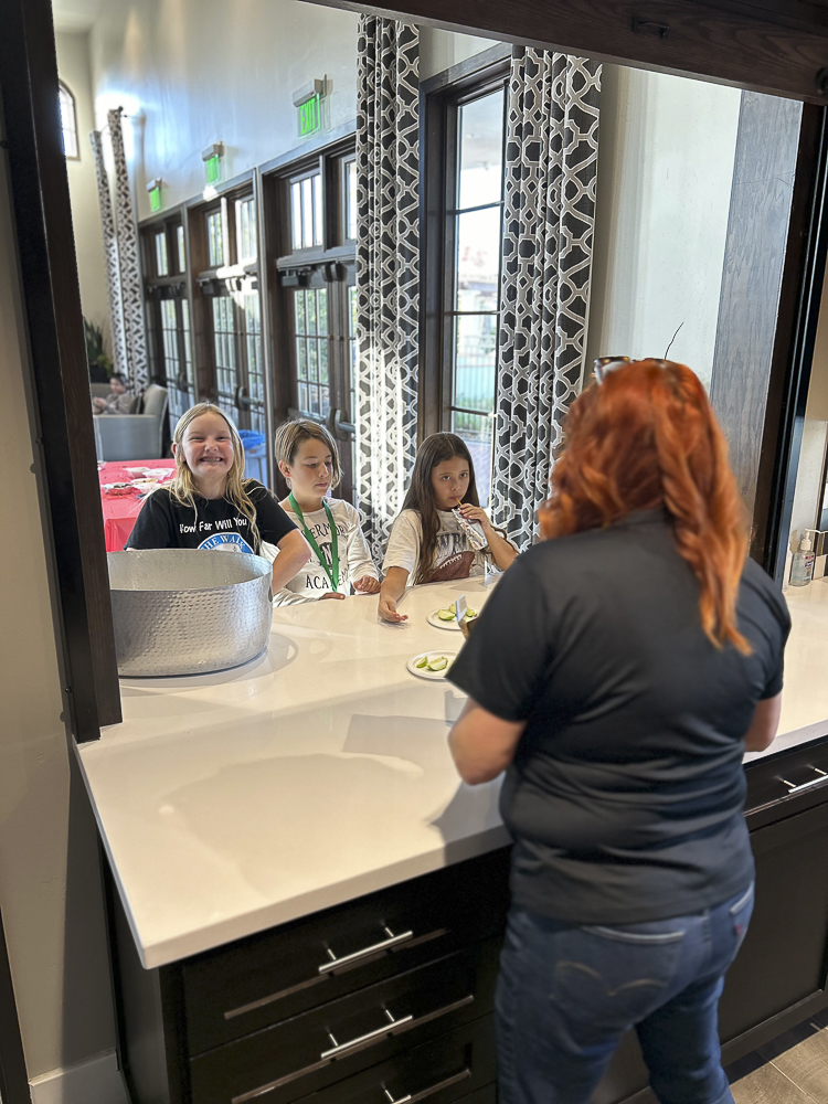 A woman with red hair serves three children at a counter. The children are eagerly reaching for cucumbers on a plate. The background features large windows and patterned curtains. The atmosphere is light and friendly.