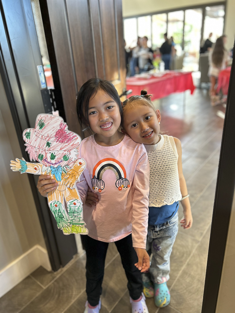 Two young girls smiling at the camera in a room with large windows. One girl holds a colorful paper cutout of a person. The other girl has a bow in her hair. Both are casually dressed and appear happy.