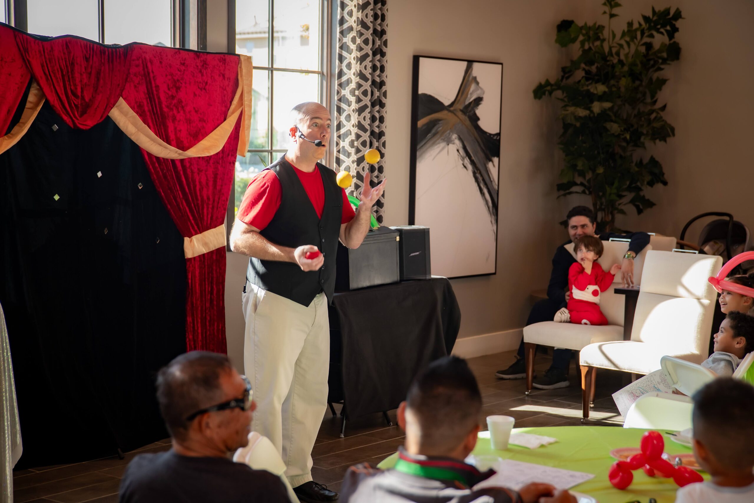 A performer juggles colorful balls in front of an audience in a decorated room. The audience, including children and adults, watches attentively. Red curtains and abstract art adorn the background.