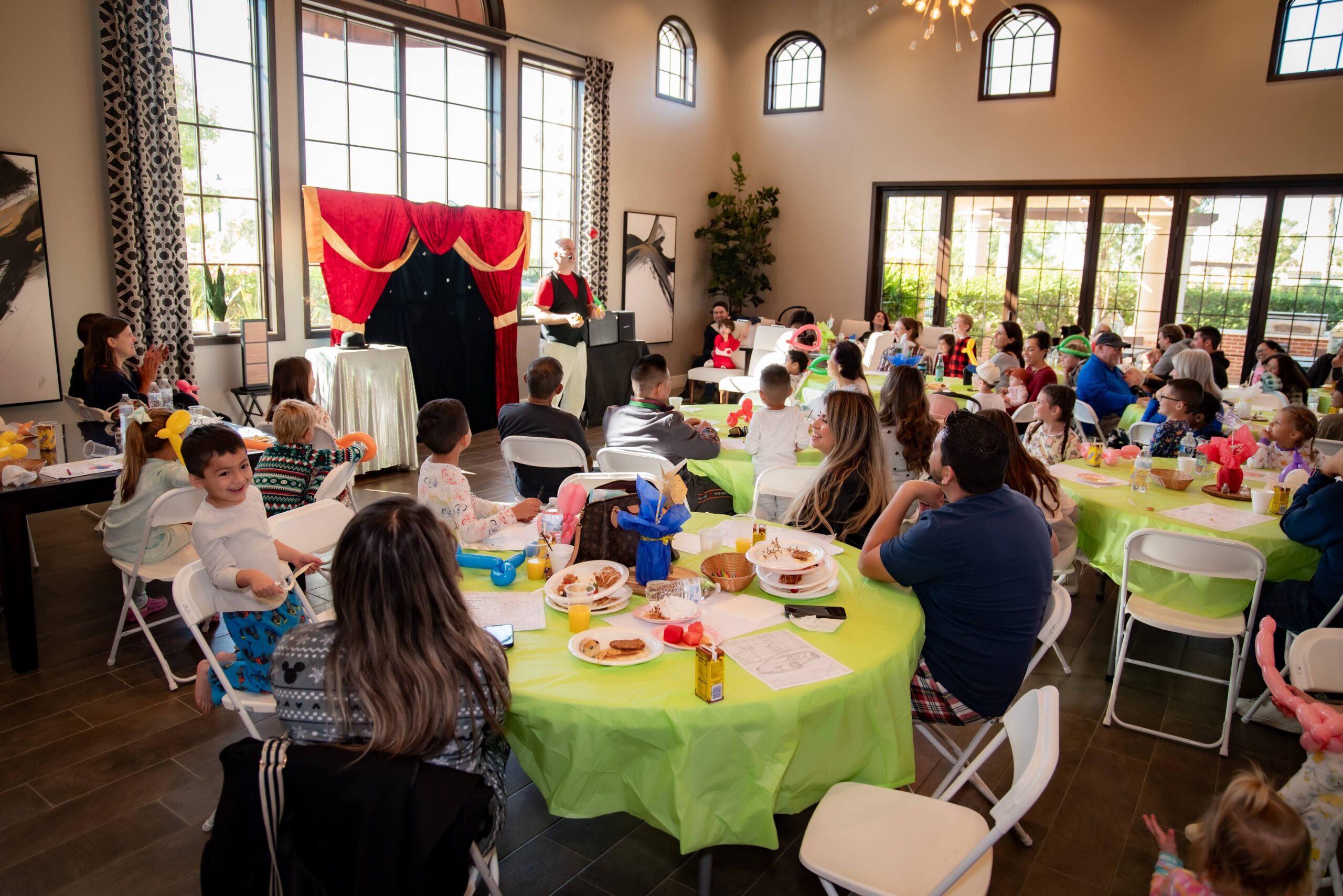 A children's party with guests seated at round tables covered in green tablecloths. A performer in front of a red curtain entertains the group. The room has large windows, letting in natural light, and decorations create a festive atmosphere.