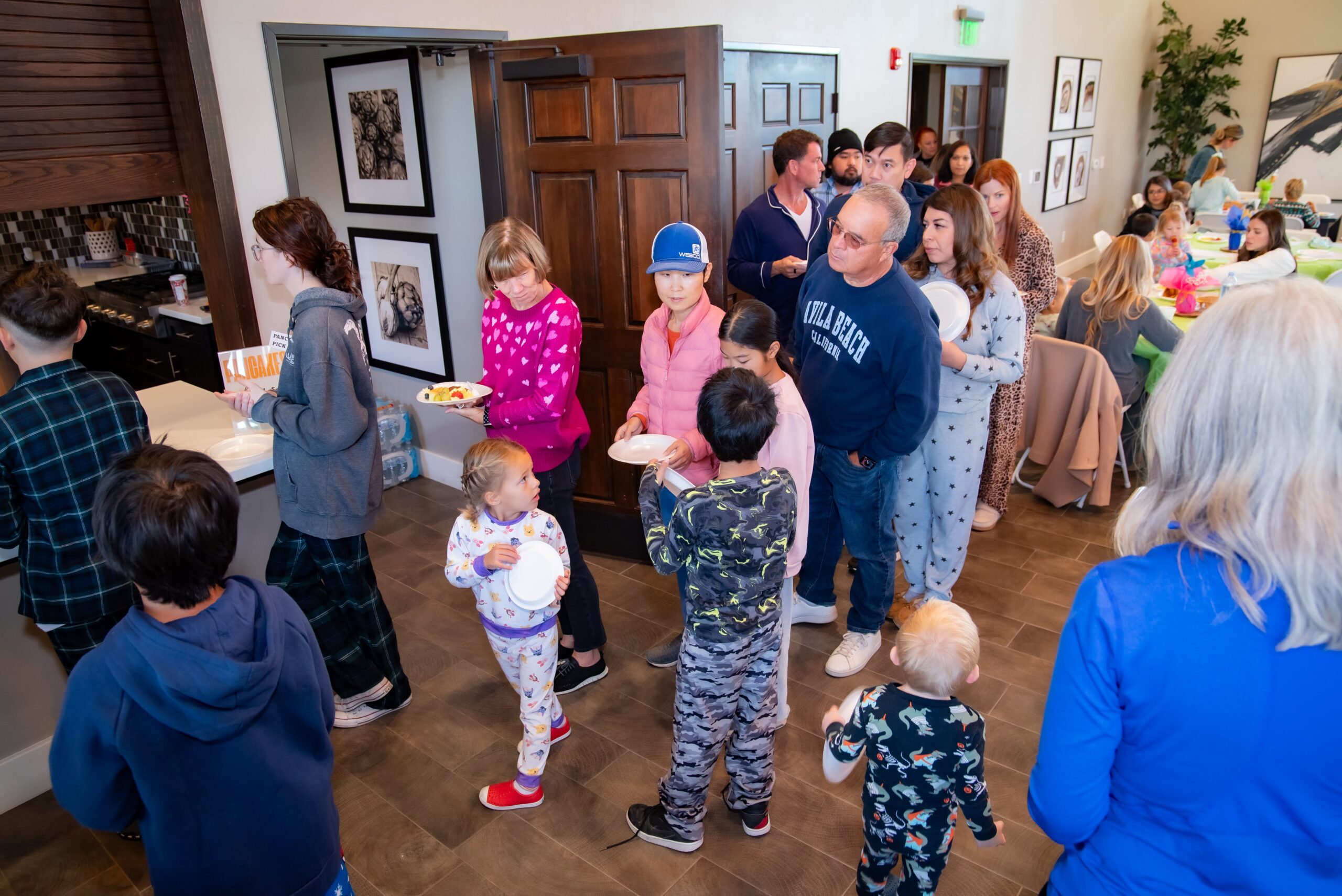 A group of people, including adults and children in pajamas, stand in line at a buffet-style gathering. Some hold plates while others chat or serve themselves. The room is bright and decorated with framed artwork on the walls.