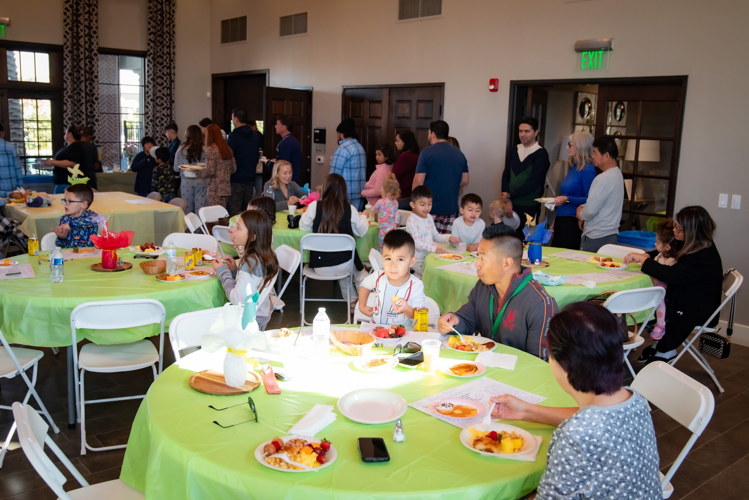 A group of people of all ages gathered in a spacious hall with round tables covered in white cloths. Some are sitting and eating, while others stand and chat. Plates and a variety of colorful dishes are visible on the tables.