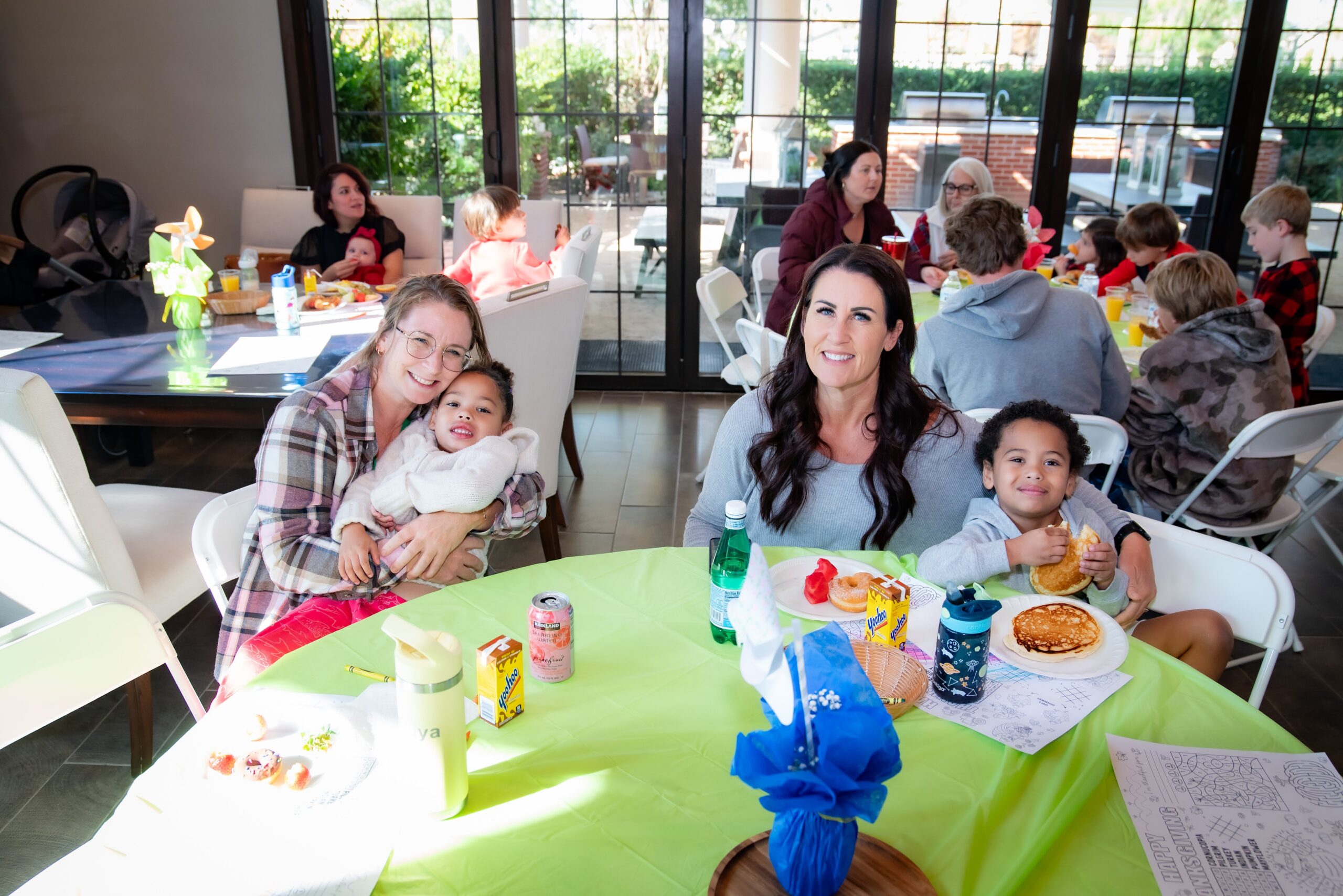 A lively family gathering with women and young children seated at a table enjoying a meal. The table is covered with a green tablecloth, filled with snacks and drinks. Large windows in the background let in natural light, and others mingle in the room.