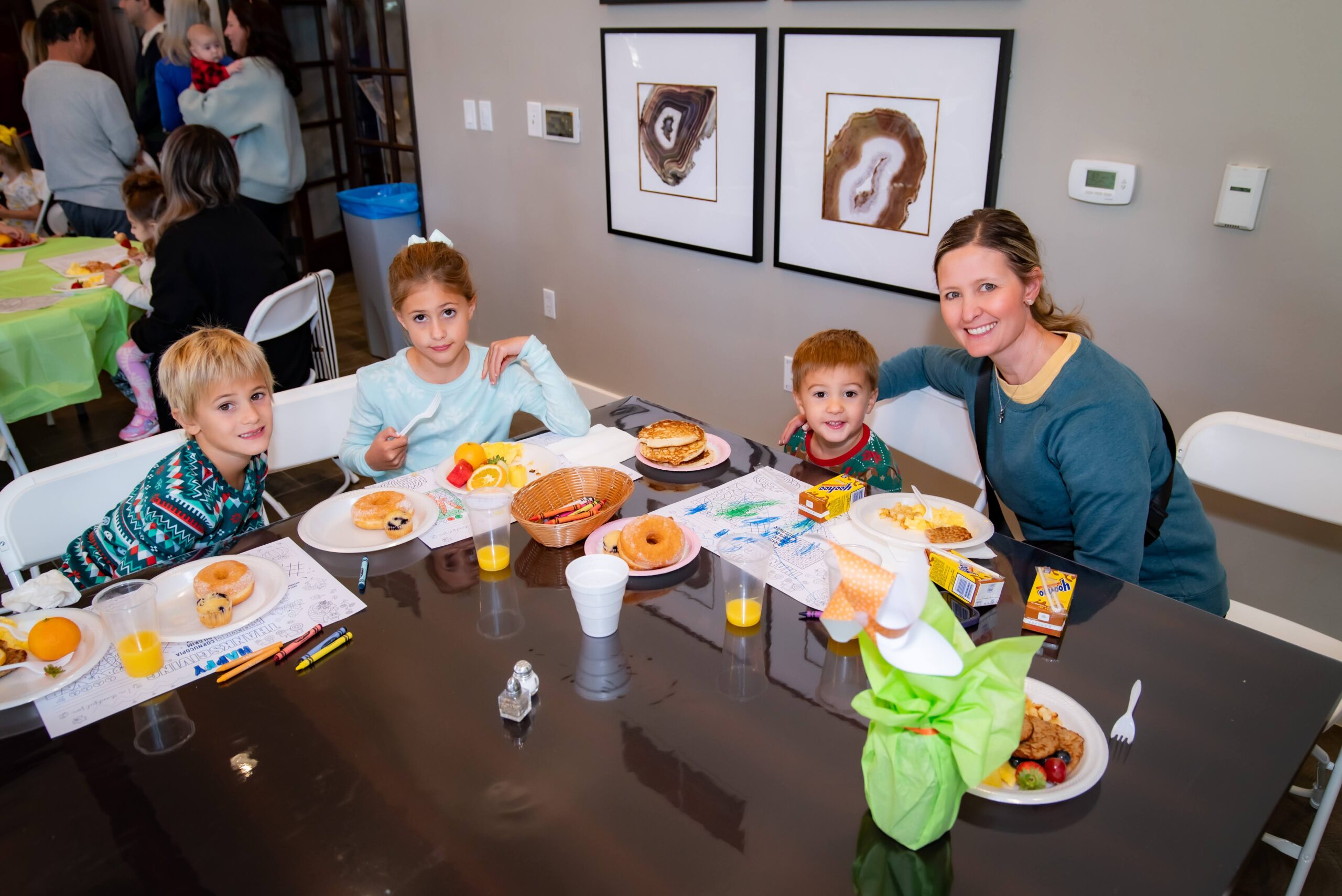 A woman and three children sit around a dining table with food and drinks, smiling at the camera. The table has breakfast items like donuts, oranges, and juice. Other people are in the background, and two framed artworks are on the wall.