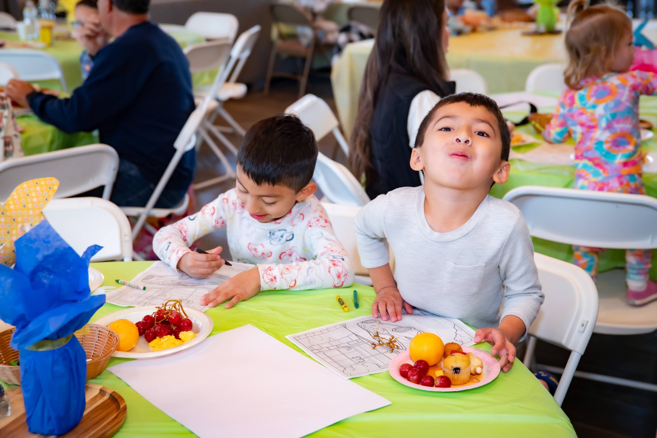 Two children sit at a table during a gathering. One child is drawing on paper, while the other makes a playful face, holding a plate with fruit and a pastry. The table is covered with a green tablecloth and has decorations. People are seen in the background.