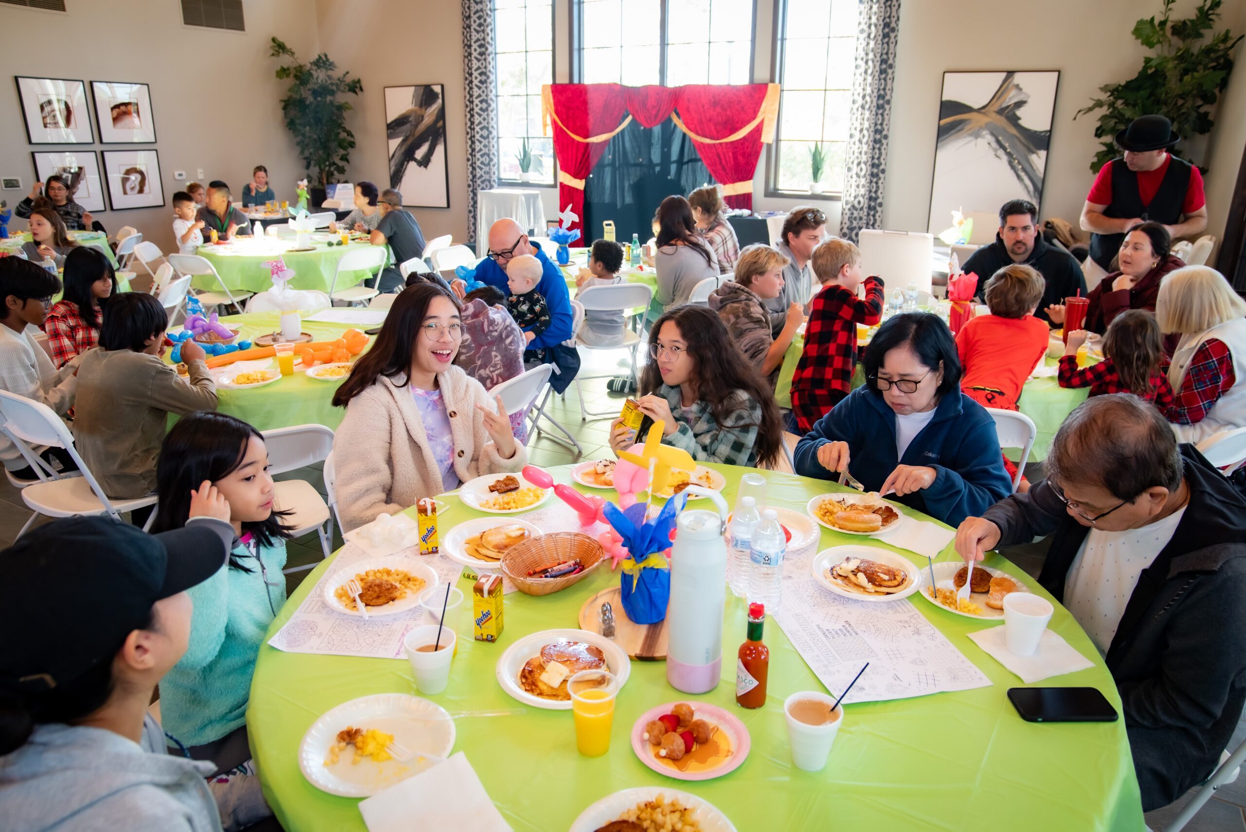 A group of people of diverse ages gather in a bright hall, enjoying breakfast at decorated tables with green tablecloths. Some converse, others eat pancakes. A stage with red curtains is in the background, indicating a festive event.