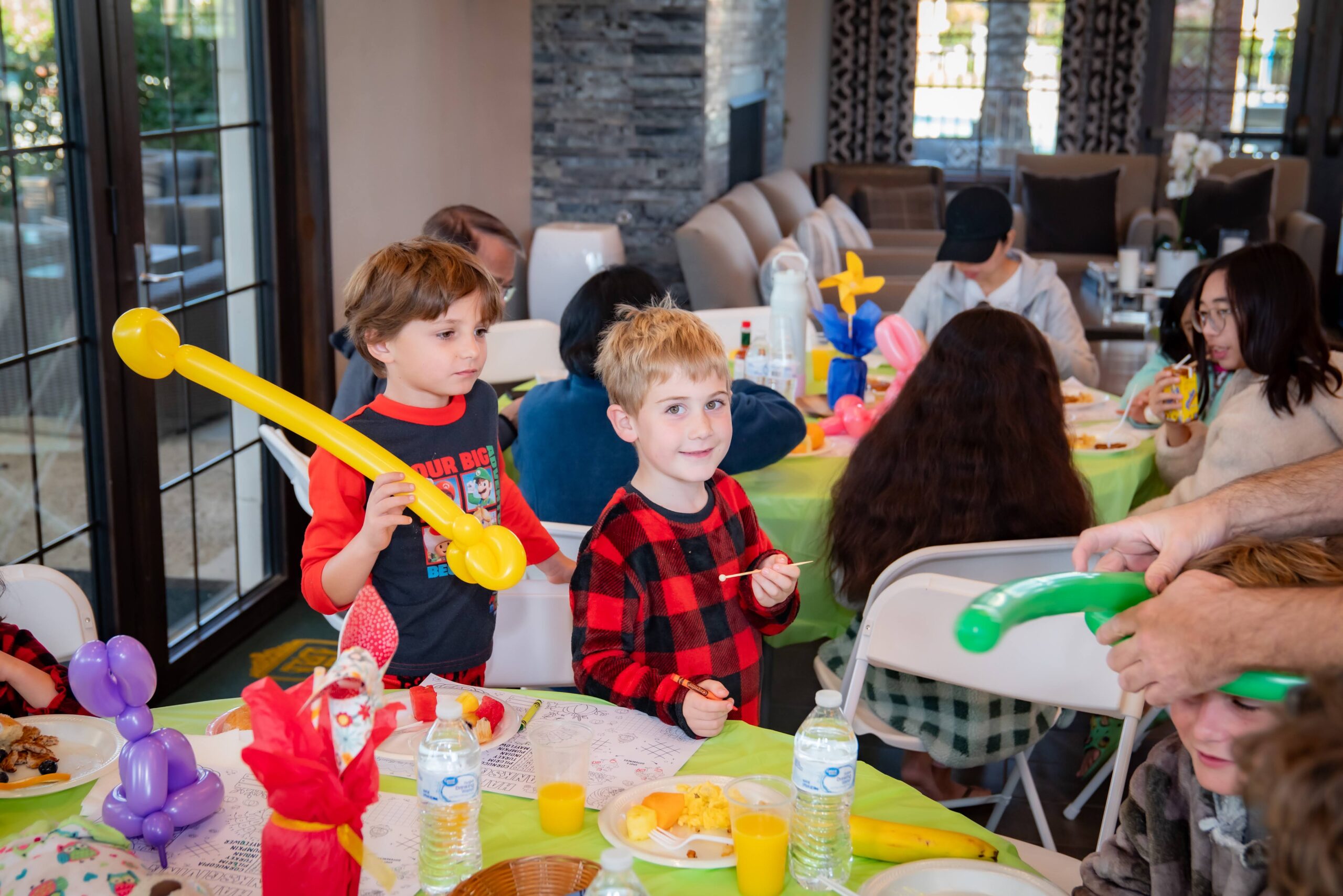 Two children in a festive setting, each holding colorful balloon animals. They stand beside a table with food and drinks, surrounded by other people seated at tables. The room has large windows and a cozy atmosphere.