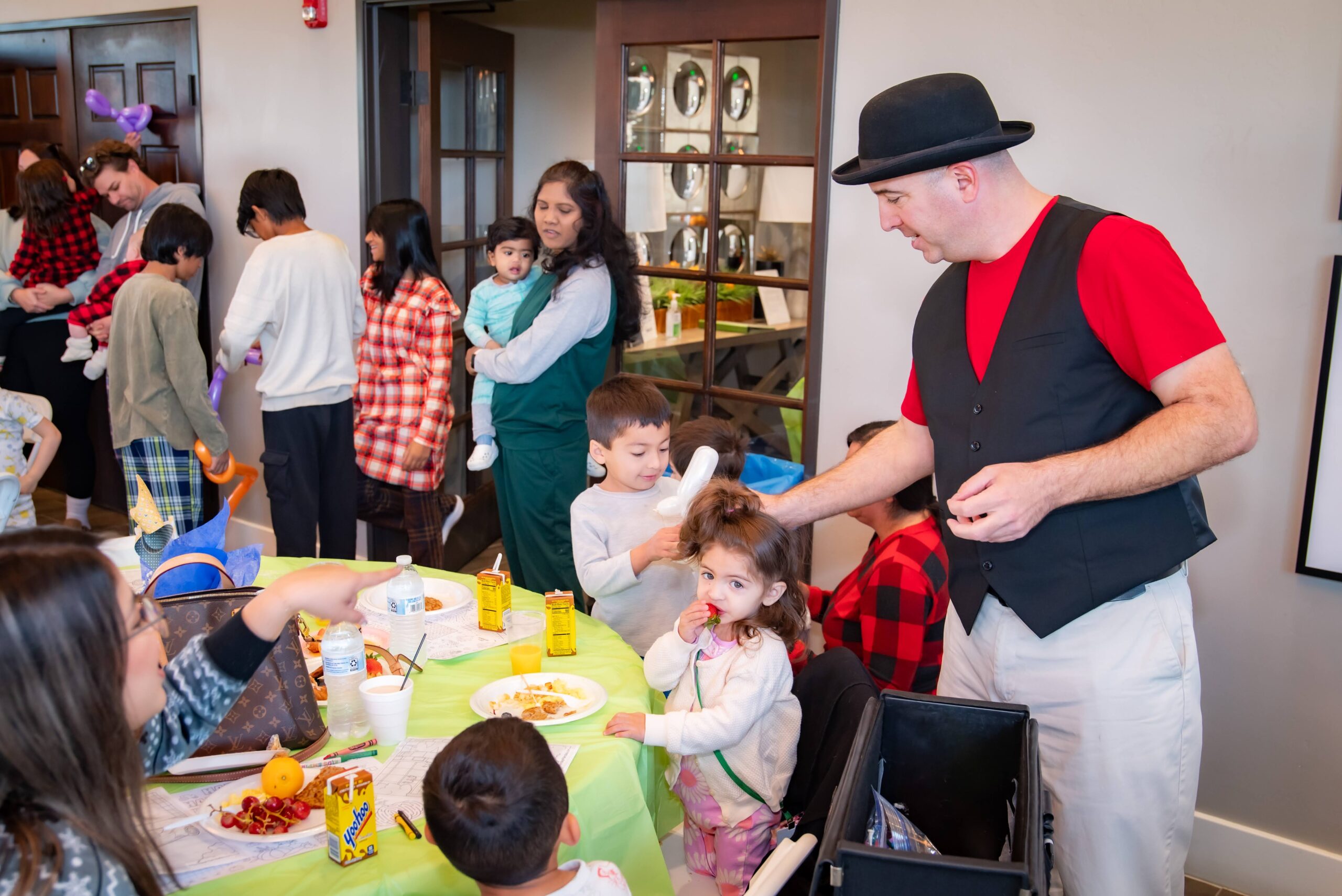 A man in a hat and vest entertains children at a party. Kids are engaged with activities, eating snacks, and wearing festive attire. A woman holds a child nearby, and tables are decorated with colorful items. It's a lively and cheerful atmosphere.