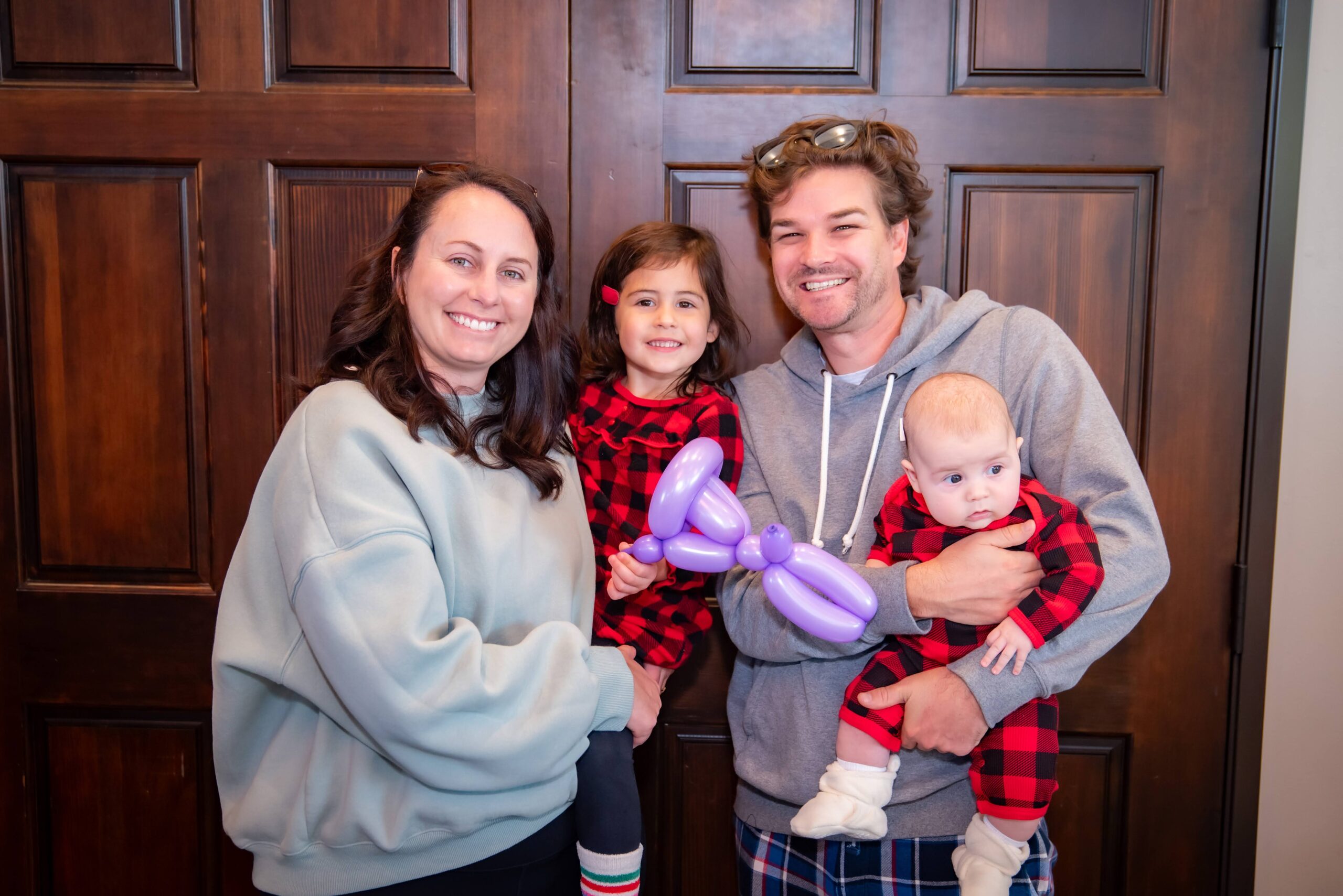 A smiling family poses in front of wooden doors. The father holds an infant and wears a gray hoodie, while the mother wears a light gray sweatshirt. A young girl in matching plaid pajamas holds a purple balloon animal.