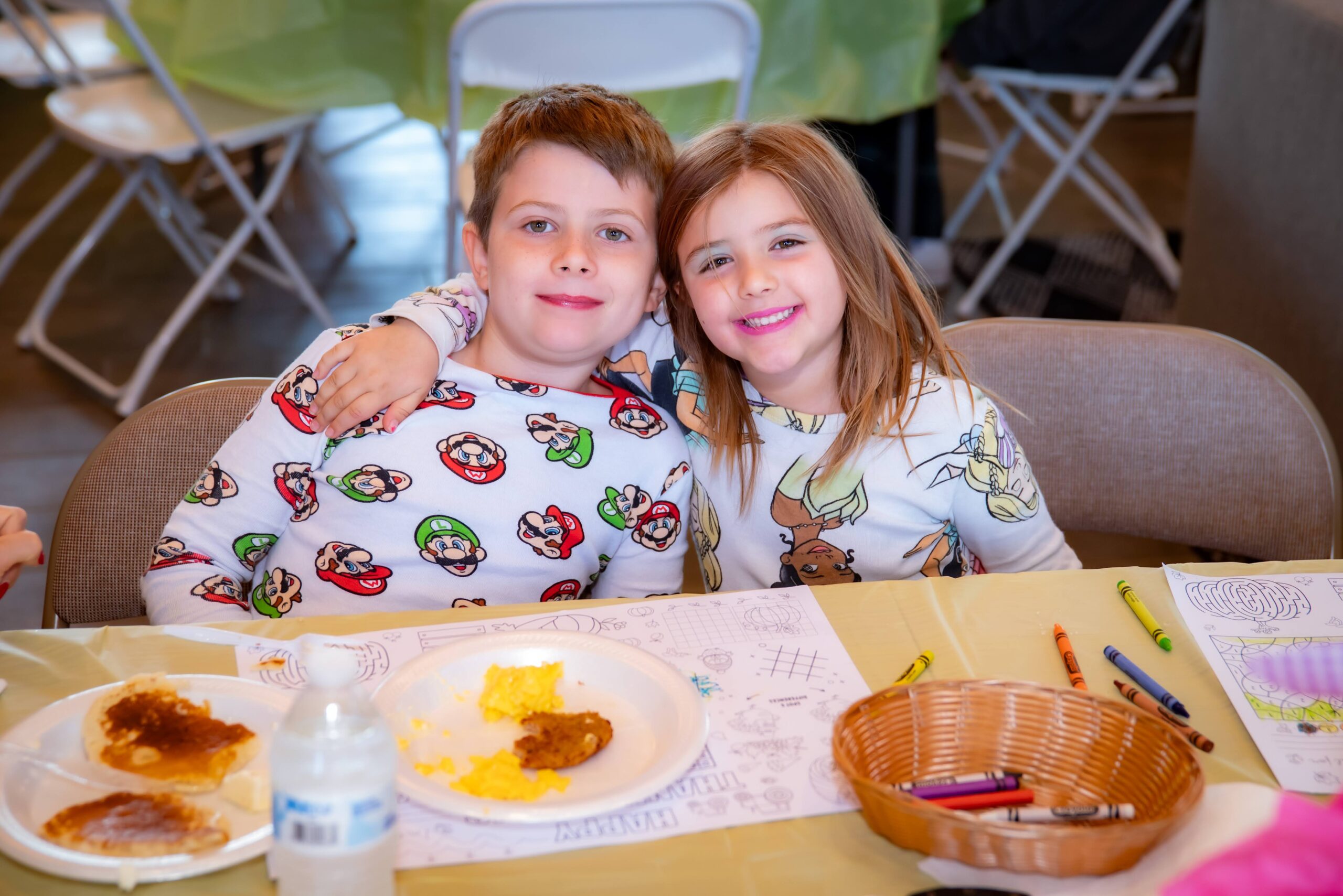 Two children sitting at a table, smiling and wearing festive pajamas. The table has plates with food, coloring pages, and crayons. They are in a bright room with folded chairs in the background.