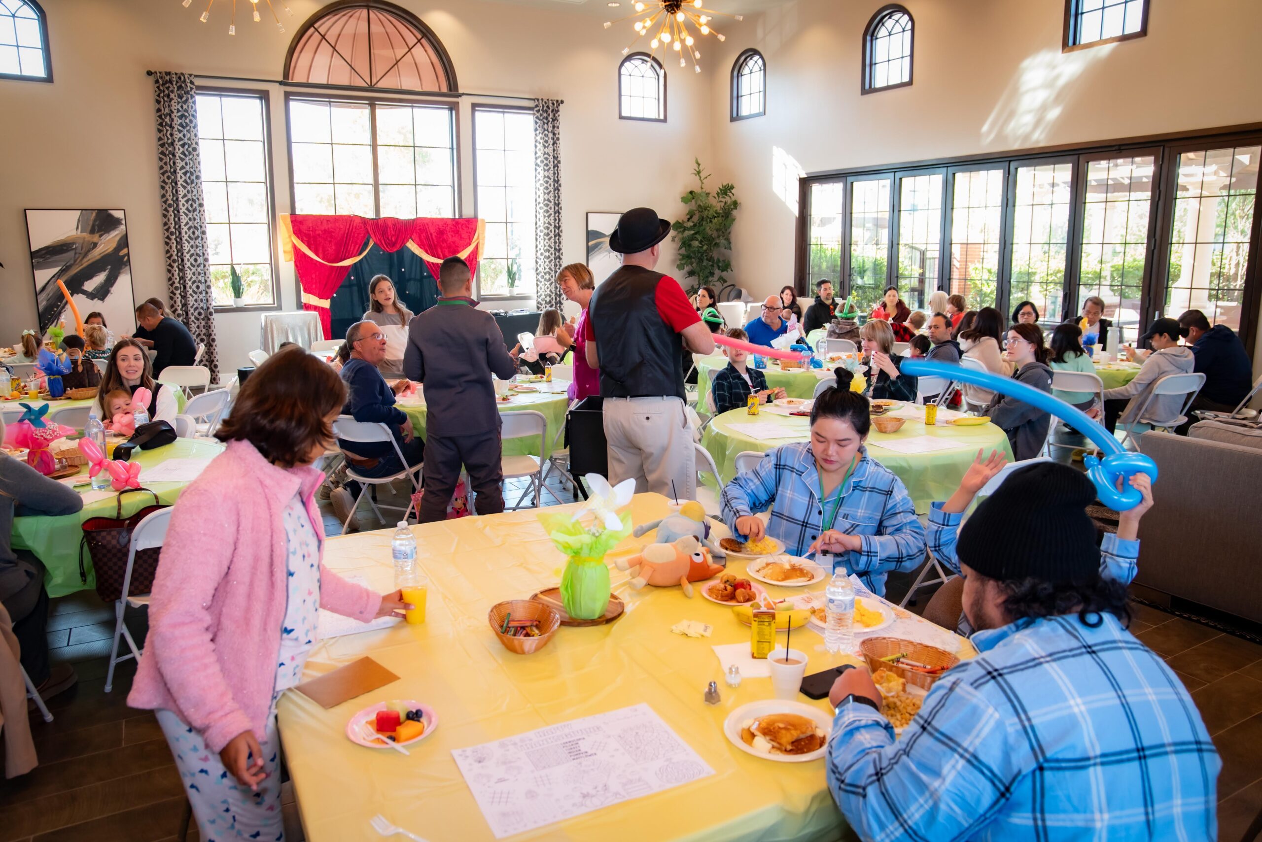 A lively indoor gathering with people seated at round tables enjoying a meal. A person in a hat entertains guests by twisting balloons. The room is bright, with large windows and festive decorations, creating a cheerful atmosphere.