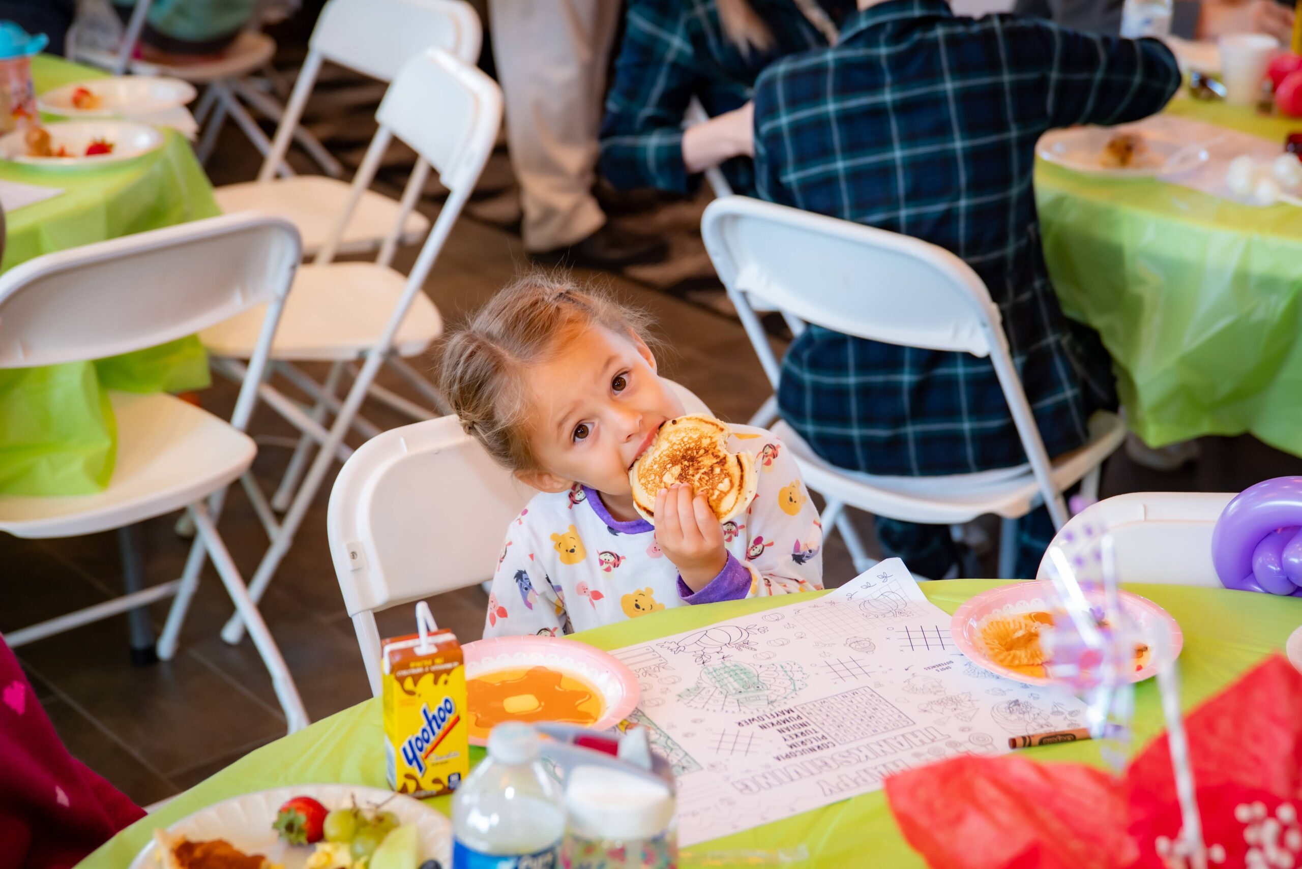 A young girl sits at a table and eats pancakes, surrounded by plates of food. She wears a colorful shirt and is seated on a white chair. Other people are visible in the background, and the table is covered with a green tablecloth.