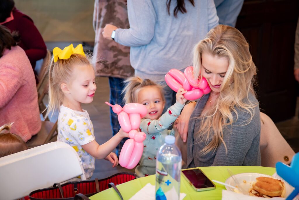 Two young children playfully present balloon animals to a smiling woman at a table. The children are joyful, and one wears a yellow bow. The table has a water bottle and a phone. Others are blurred in the background.