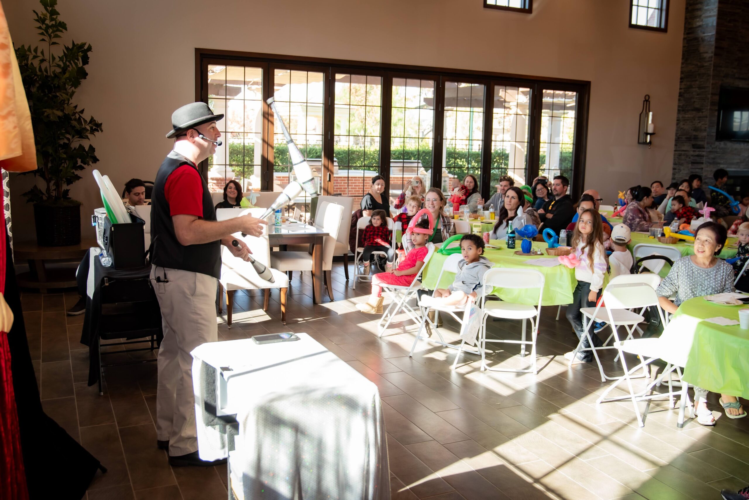 A performer, wearing a hat and headset, entertains a group of adults and children seated at tables covered with green tablecloths in a sunny room with large windows. The audience appears engaged and attentive.