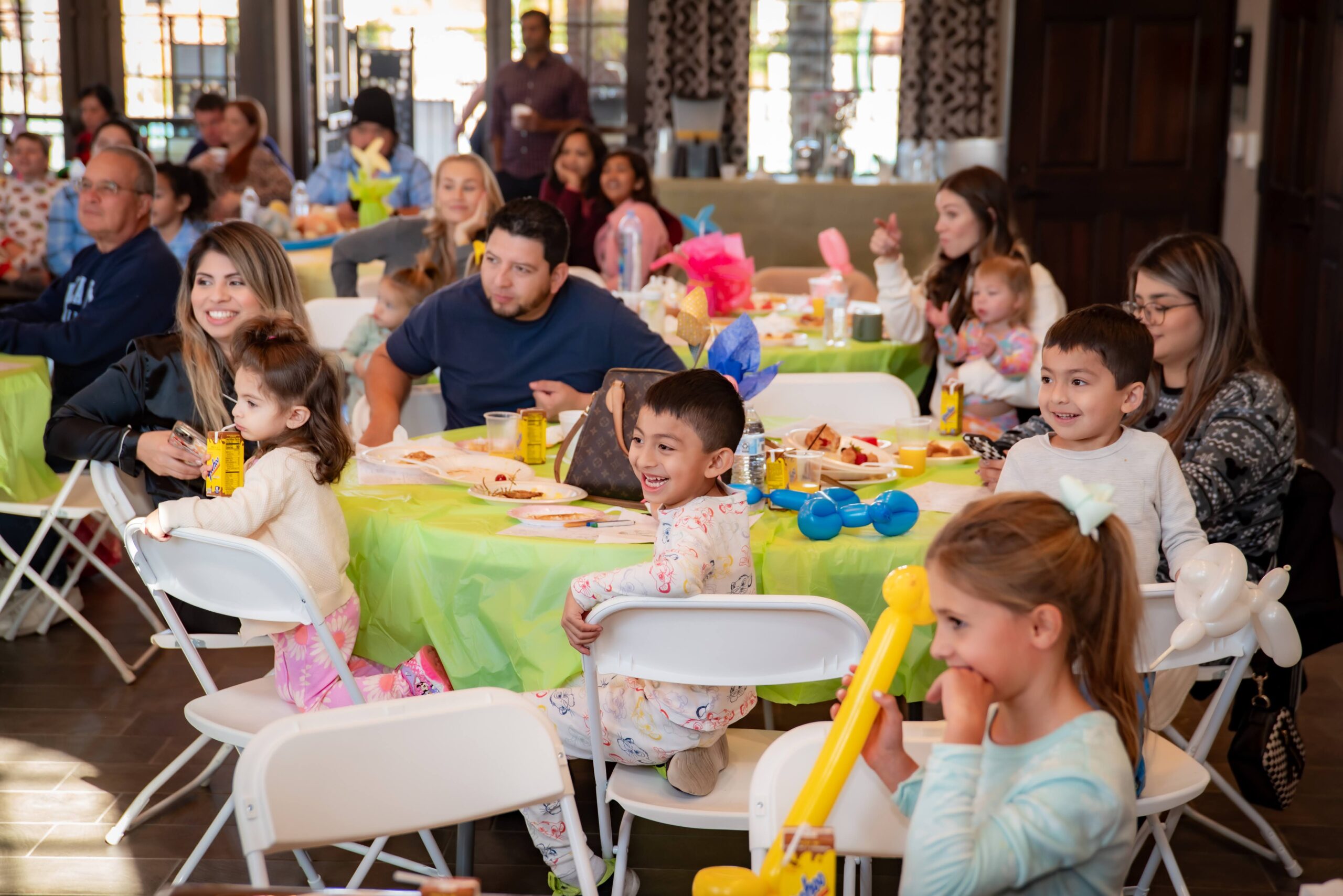 A group of adults and children enjoy a festive gathering, seated at tables with green tablecloths. The children appear entertained, holding balloon animals. Bright decorations suggest a joyful atmosphere indoors.