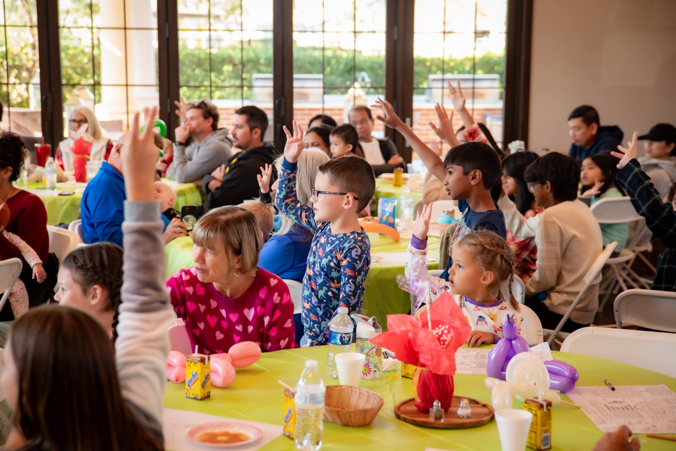 Children and adults sitting at tables with colorful tablecloths, raising their hands enthusiastically. The room is bright with natural light from large windows in the background.