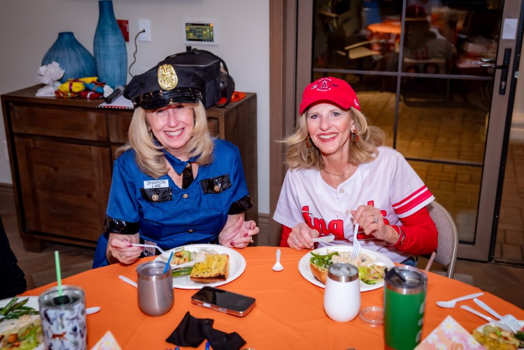 Two women sitting at a table, wearing costumes. One is dressed as a police officer, and the other in a sports uniform. They are smiling and eating food from plates. The table has orange and yellow decorations.