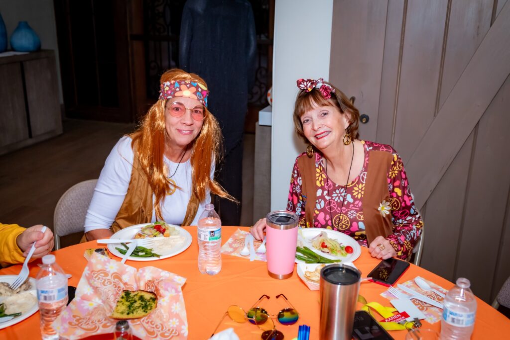 Two women dressed in retro hippie outfits sit at a table with plates of food. The table is covered with an orange tablecloth and has bottles of water and a pink tumbler. The women are smiling and wearing headbands and colorful dresses.