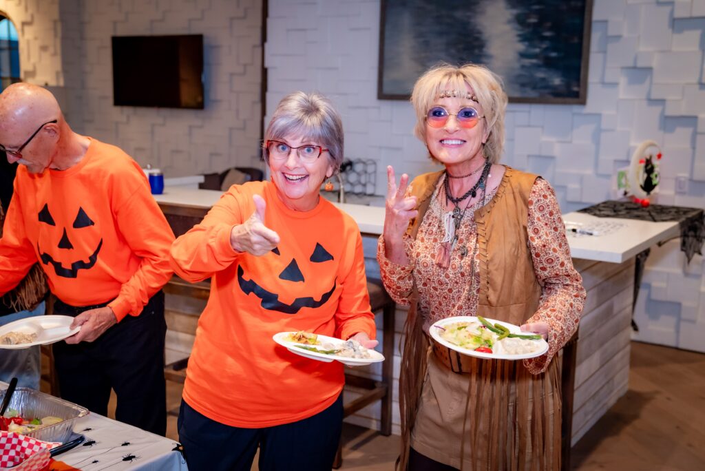 Two women smiling at a Halloween-themed party. One is wearing an orange shirt with a jack-o'-lantern face, and the other is in a brown dress with glasses. They are holding plates of food, with a man serving himself in the background.