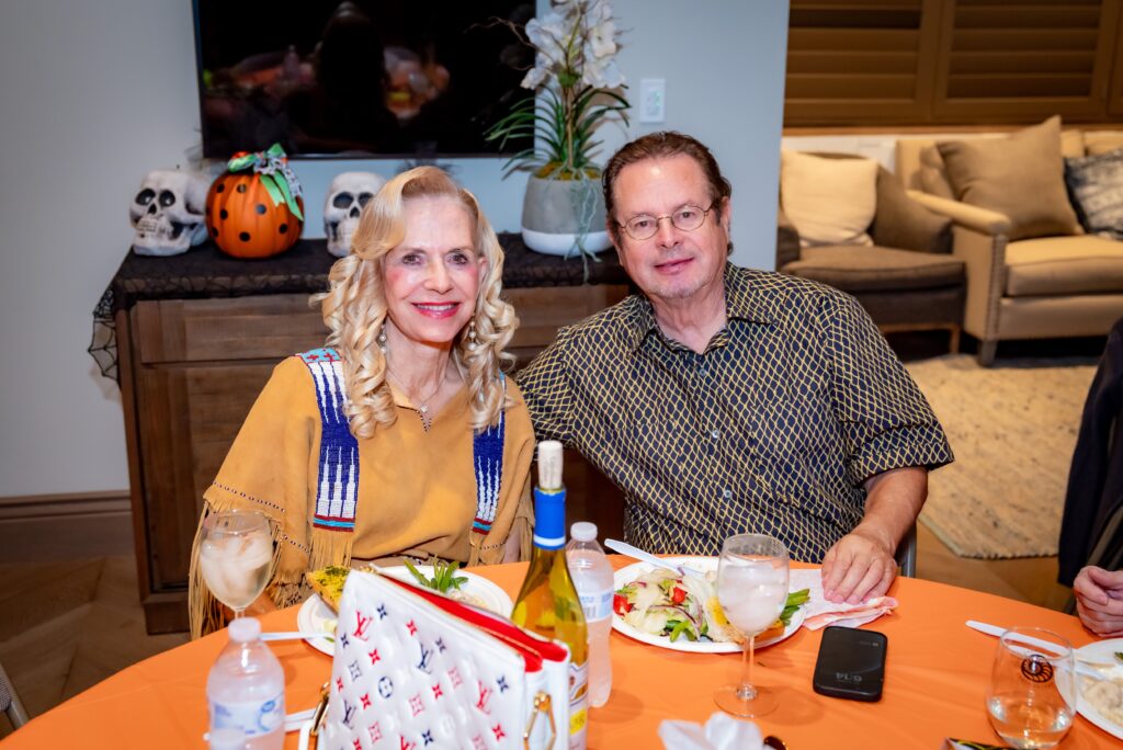 A smiling couple sits at a table with dinner plates, surrounded by Halloween decorations, including skulls and a pumpkin. The woman wears a tan outfit with blue accents, and the man wears a patterned shirt.