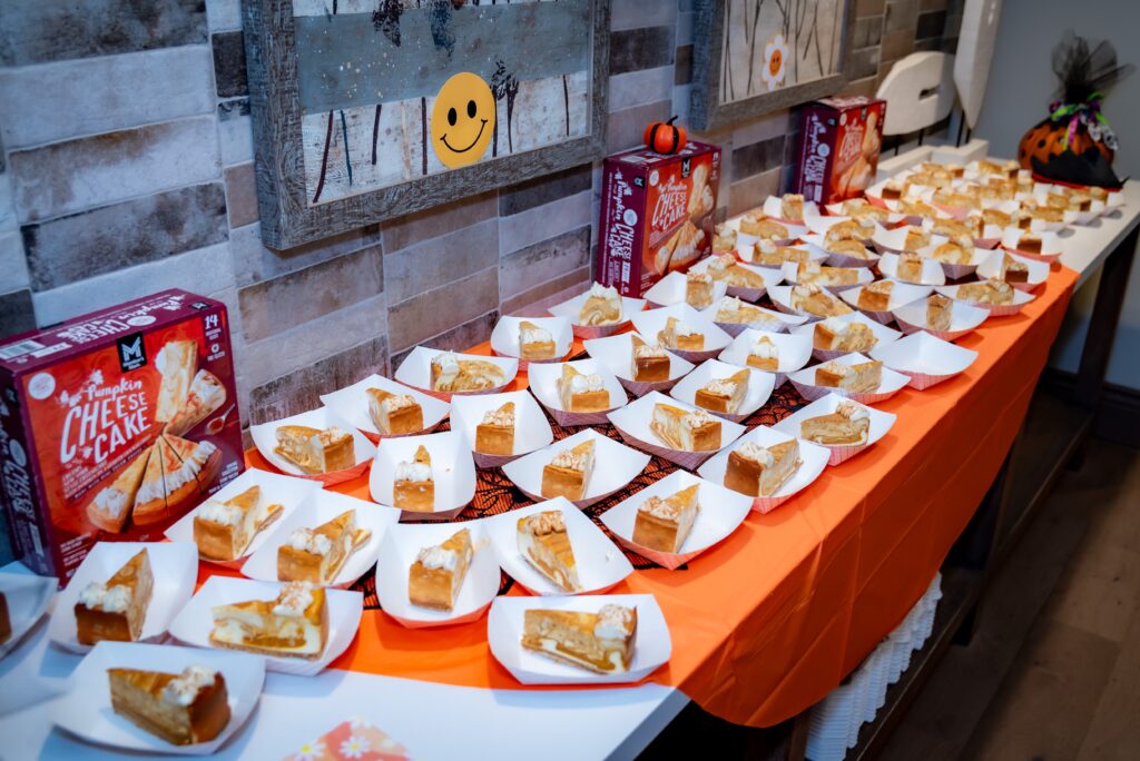 A table is covered with numerous plates, each holding a slice of pumpkin cheesecake. The table is decorated with orange tablecloths, and Halloween-themed boxes and decorations are visible in the background.