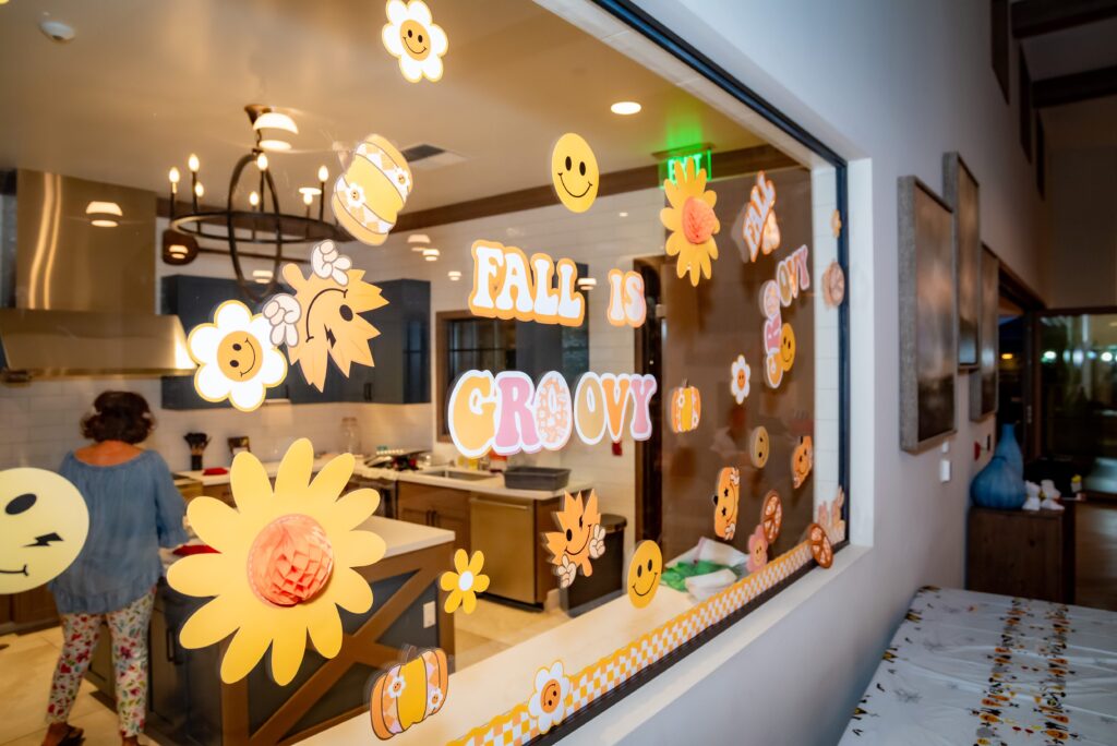 A window display with floral and smiley face decals and the words "Fall is Groovy" in colorful letters. Behind the window is a kitchen with a chandelier and a person standing inside. A table with a patterned cloth is nearby.