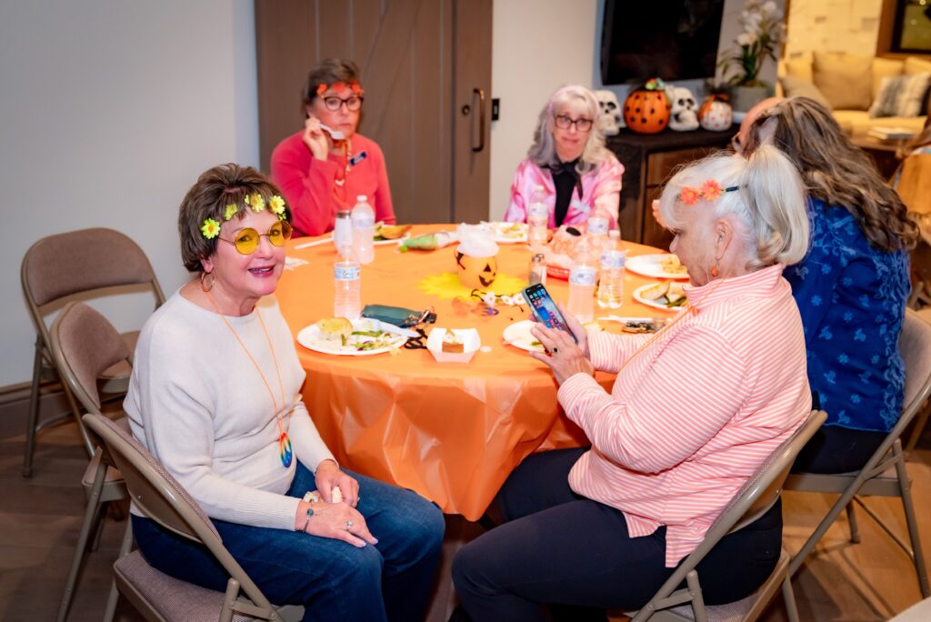 Five women are seated around a table decorated with an orange tablecloth, eating and chatting. One woman wears yellow sunglasses and a flower crown, while another checks her phone. The room is decorated with fall-themed items like pumpkins.