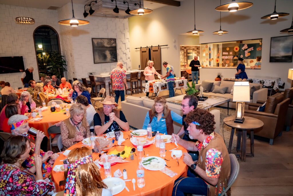 People in colorful outfits are gathered indoors for a festive meal. Tables are decorated with orange tablecloths, and attendees are dressed in 70s-style attire. The background features Halloween-themed decorations.