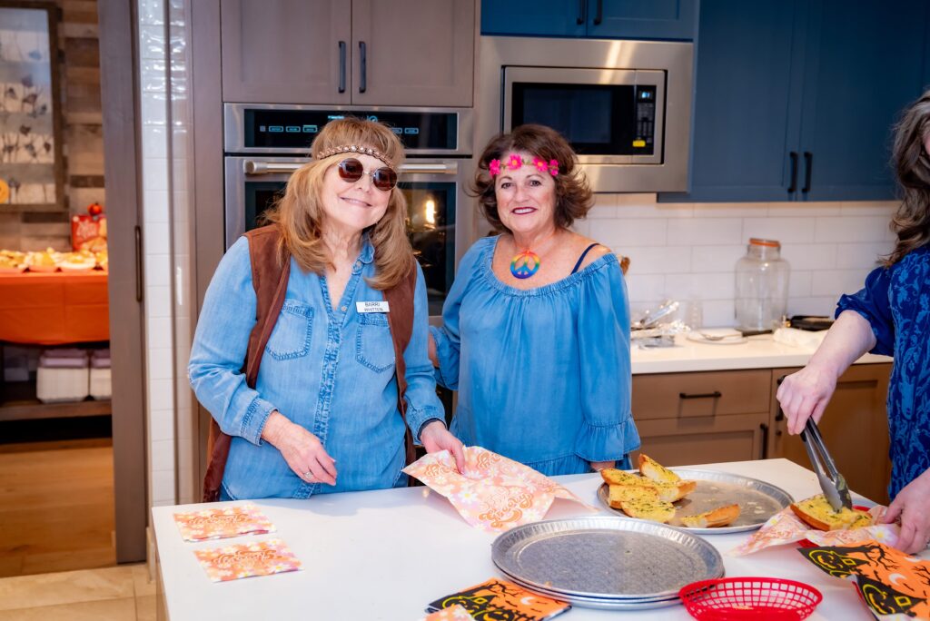 Two women in a kitchen wearing denim outfits and sunglasses are serving garlic bread onto plates. One has a pink flower headband, and there are autumn-themed napkins on the counter.