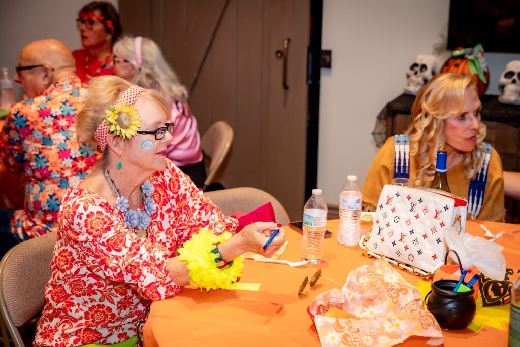 A lively party setting with colorful decor. Guests, dressed in vibrant costumes and accessories, are seated around tables with bright orange tablecloths. Skulls and festive items adorn the room, creating a fun and eclectic atmosphere.