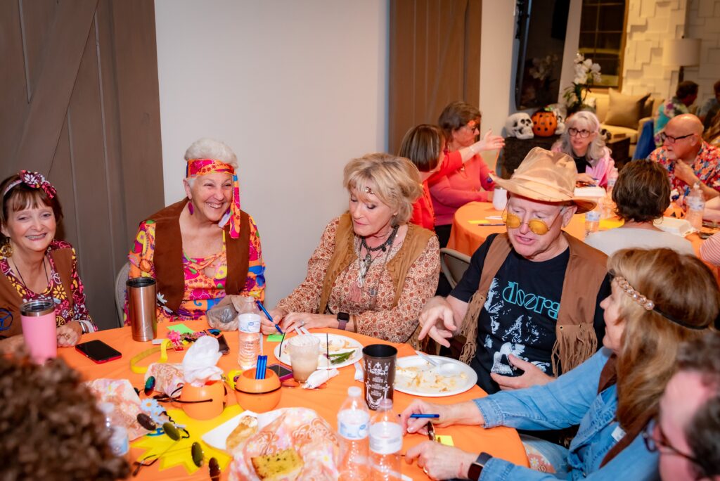 A group of people in colorful, retro costumes enjoying a meal at a table decorated for Halloween. The room is vibrant with festive attire and background details, adding to the lively atmosphere.