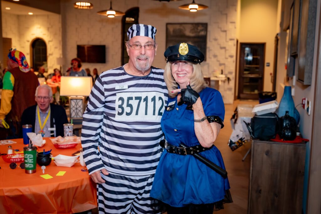 A man in a prisoner costume with black and white stripes stands next to a woman dressed as a police officer. They are posing and smiling at a party, with other guests and decorations visible in the background.