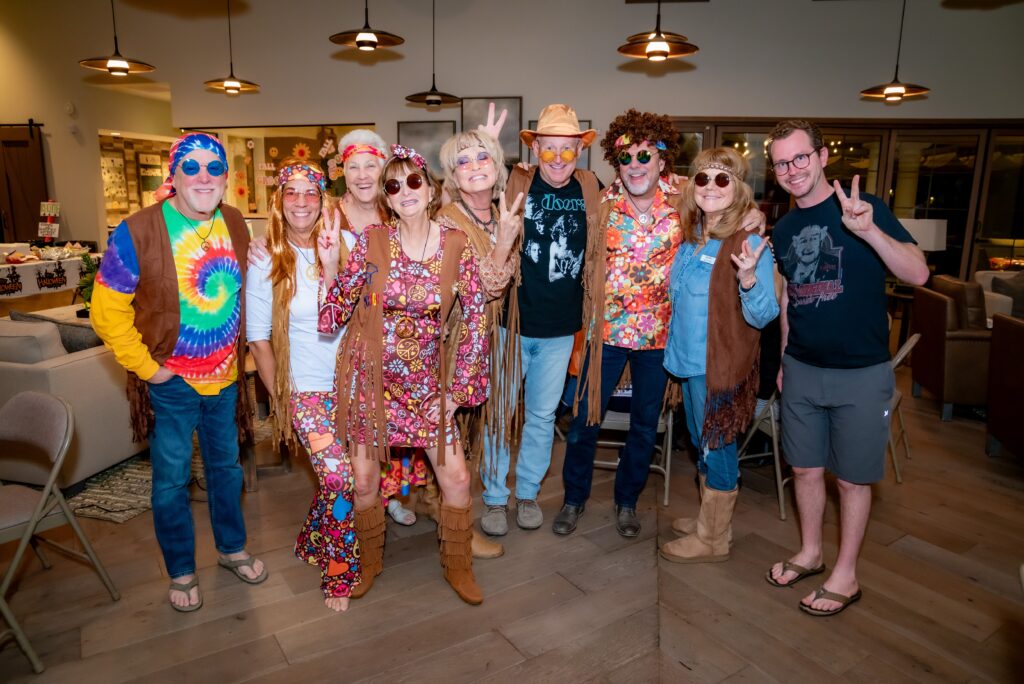 A group of people wearing colorful, retro costumes reminiscent of the 1960s, with tie-dye, floral patterns, and peace signs. They stand indoors, smiling and posing playfully for the camera.