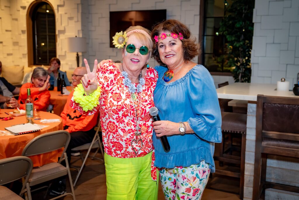 Two women in colorful outfits, one with green sunglasses and peace sign, the other holding a microphone, stand smiling at a lively indoor gathering. People in the background are seated at tables.