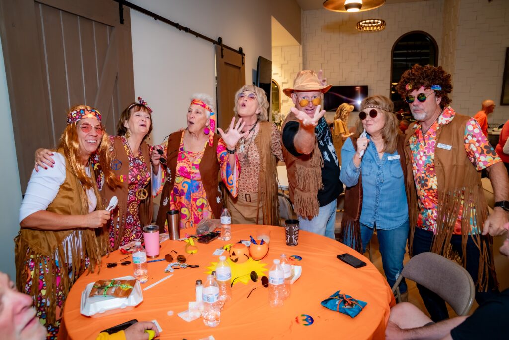 A group of people dressed in colorful 70s-themed costumes, including fringe vests and floral patterns, gather around an orange table. They are posing cheerfully, some making peace signs and wearing sunglasses or hats.