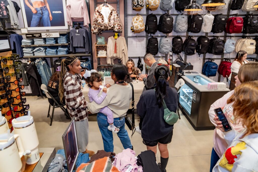 A crowded clothing store with people browsing. Backpacks hang on the wall, and sunglasses are displayed on a counter. A person holds a baby, while others look at merchandise. Shoppers are interacting with the store environment.