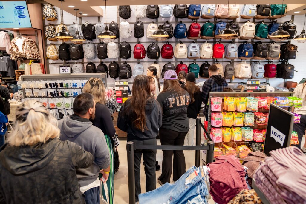 Shoppers stand in line inside a store, surrounded by shelves of colorful backpacks and racks filled with clothes. A wall display of snacks is visible on the right. The scene is busy and vibrant, suggesting a sale or popular shopping event.