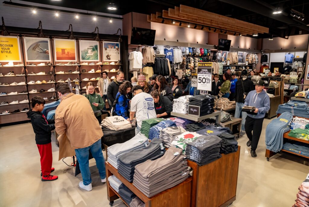 A busy clothing store interior with numerous shoppers browsing racks and shelves filled with folded clothes. A sign advertises a 50% off sale. The atmosphere is lively with people of various ages interacting and shopping.