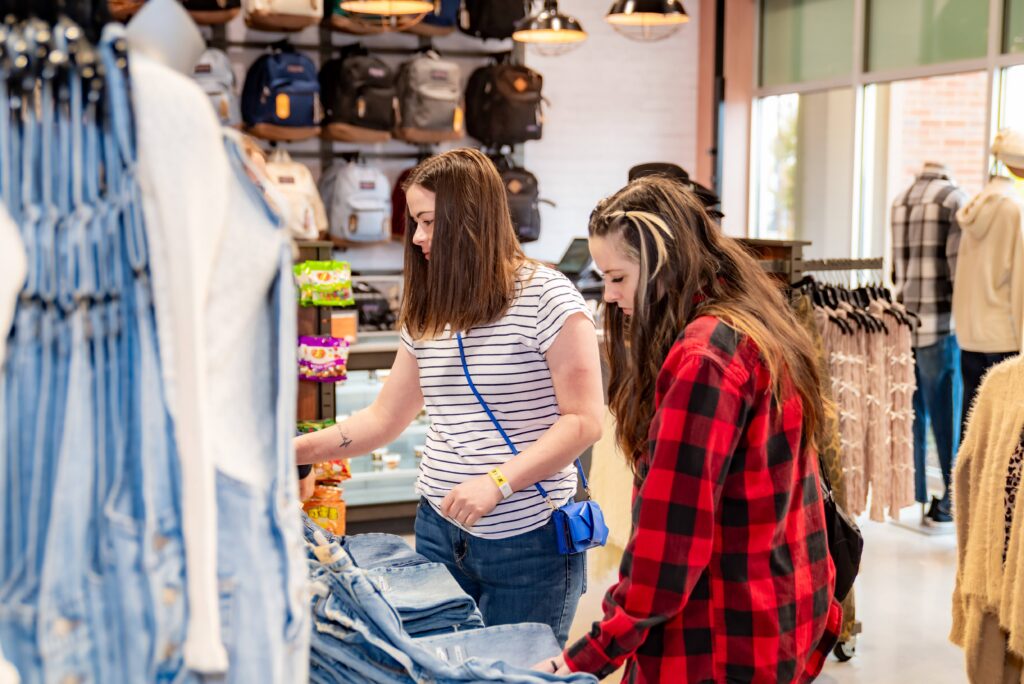 Two women shop for denim jeans in a store. One wears a striped shirt and blue crossbody bag, while the other wears a red plaid shirt. In the background, bags and clothing are on display. Natural light enters through large windows.