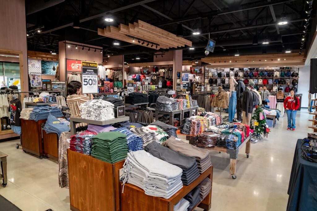 A clothing store interior filled with various folded shirts, sweaters, and jackets on display tables and racks. A sign advertises a 50% off sale. Shoppers are browsing the merchandise in a spacious, well-lit environment.