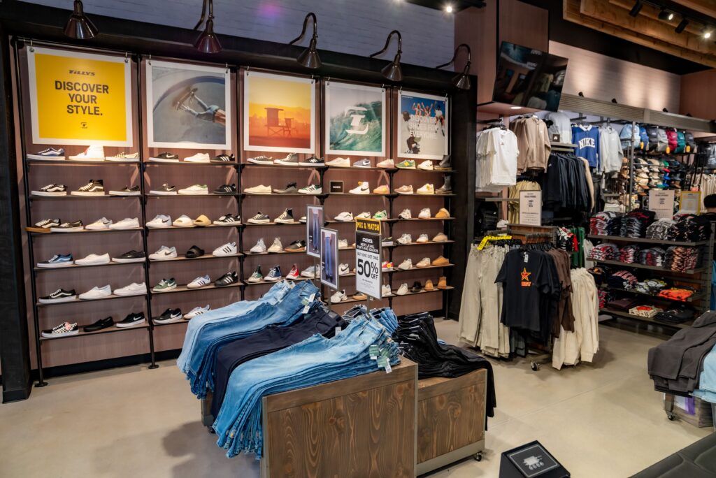 A retail store interior showcasing a wall of shoes under framed photos. In the foreground, tables display neatly folded jeans. To the right, racks hold various clothes, including hoodies and t-shirts. A sign indicates a 50% sale.