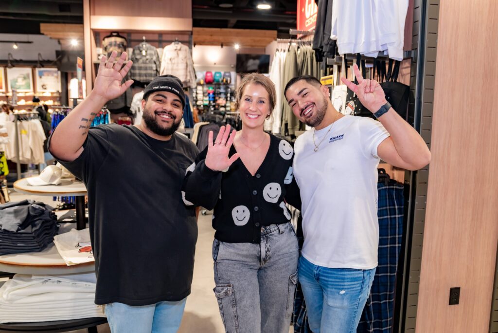 Three people smiling and standing inside a clothing store. The person on the left wears a cap and black shirt, the middle person wears a black sweater with smiley faces, and the person on the right wears a white shirt showing a peace sign.