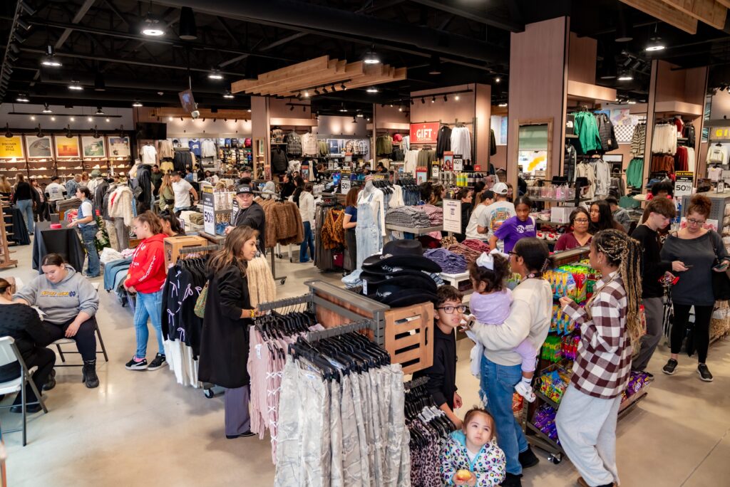 A busy clothing store filled with shoppers browsing racks of clothes. People of various ages are shopping, some with children. The store is well-lit and bustling with activity, with colorful garments displayed throughout.