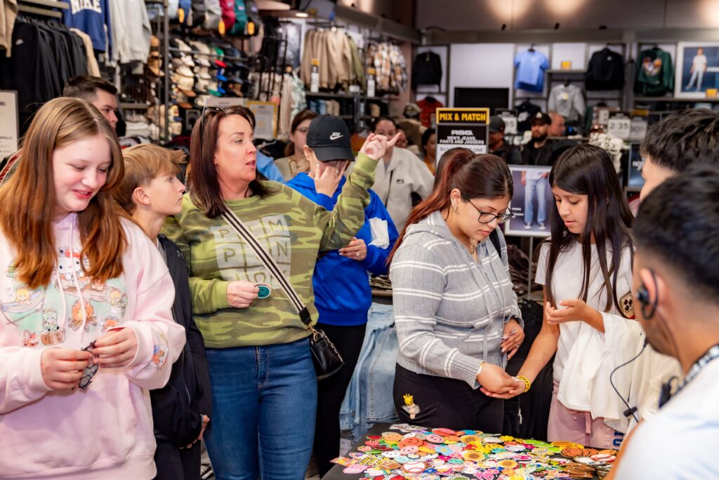 A group of people in a store browses a table filled with colorful patches and pins. A woman in a green sweater gestures, while others examine the items. The background shows shelves with clothing and accessories.