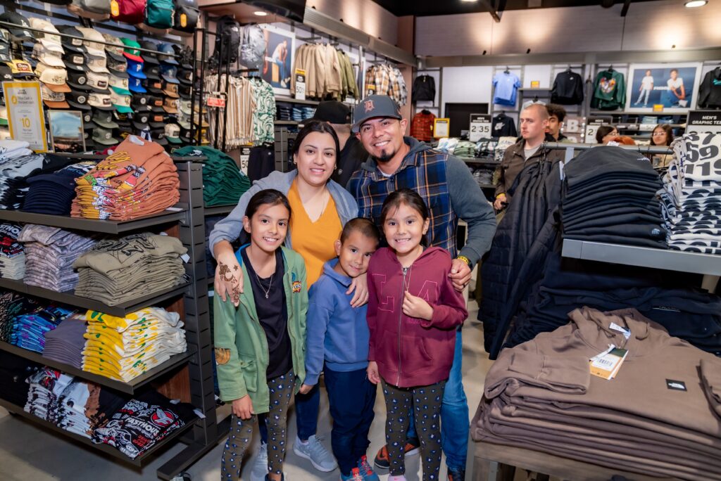 A family of five poses happily in a clothing store. The parents stand behind their three children, who are smiling. The store is filled with neatly arranged clothes and caps. Shoppers can be seen in the background.