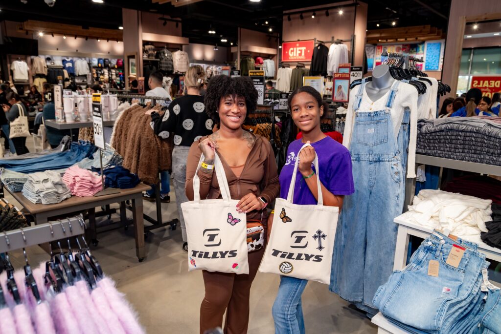 Two women smiling and holding tote bags in a clothing store. The store is filled with various apparel items like jeans and jackets displayed on racks and tables. The environment is bright, with other shoppers visible in the background.