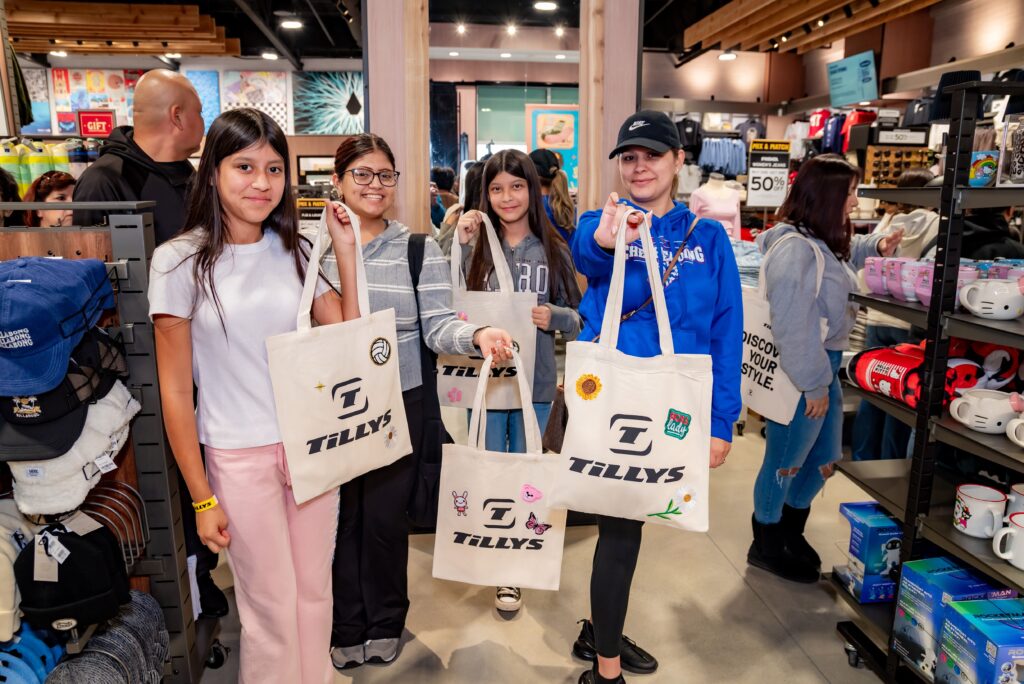 A group of four young women smile while holding personalized Tillys tote bags inside a store. Shelves with clothing and accessories can be seen around them, and other shoppers browse in the background.